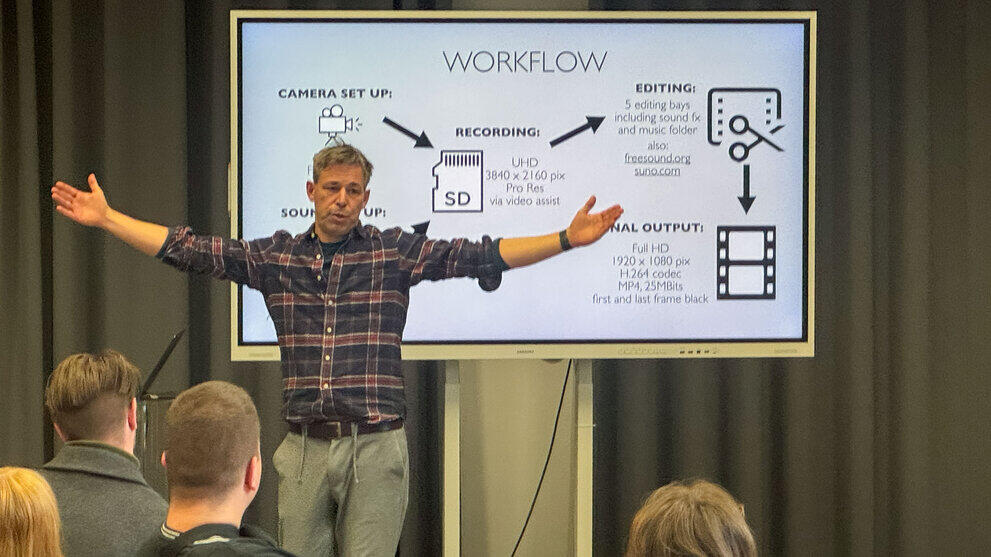 Nähere Einsstellung auf den Dozenten. Auf dem Monitor hinter ihm ist ein mit "Workflow" überschriebenes Schaubild zu sehen.